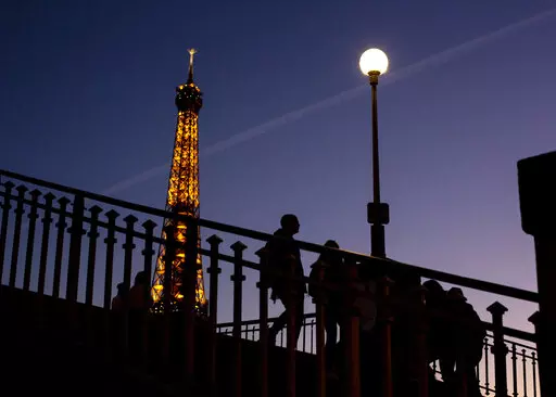 People walk on a bridge next to the Eiffel Tower in Paris, Wednesday Feb. 9, 2022. Lights on the Eiffel Tower will soon be turned off an hour earlier at night as part of an energy savings plan in the French capital, its mayor announced. Paris mayor said the iconic tower that is illuminated until 1:00am is only one of the city's monuments and municipal buildings that will be plunged into darkness earlier in the evening as the French capital faces risks of power shortages, rationing and blackouts 