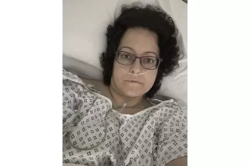 This July 2023 self-portrait provided by Anna Gittins shows the elementary school principal from Hereford in western England in a hospital bed during her cancer treatment. Gittins was shocked when she found out she had advanced colorectal cancer in 2022. But when she contacted her local hospital, she was told no one would be able to see her for three months "due to high demand and low capacity of senior doctors." Gittins had access to private health care and has since undergone surgery and chemo