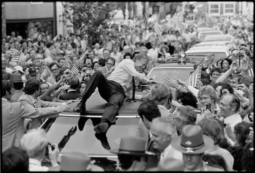 President Jimmy Carter leans across the roof of his car to shake hands along the parade route through Bardstown, Ky., July 31, 1979. The president climbed on top of the car as the parade moved toward the high school gym, where a town meeting was held. (AP Photo/Bob Daugherty, File)