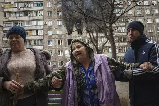 People help an elderly woman to walk in a street with an apartment building hit by shelling in the background in Mariupol, Ukraine, Monday, March 7, 2022. (AP Photo/Evgeniy Maloletka)