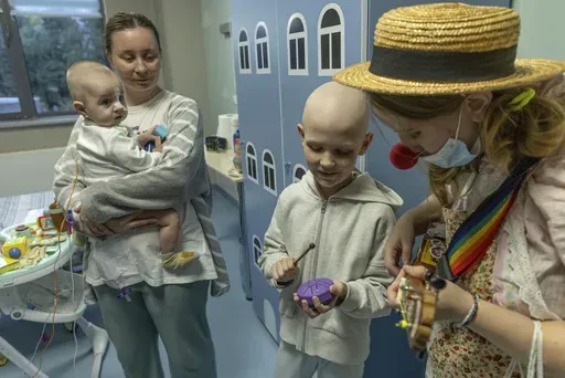 Tetiana Nosova, who goes by the clown name of Zhuzha, a volunteer from the "Bureau of Smiles and Support" plays a ukulele as she stands with Michael Bilyk, who is held by his mother Antonina Malyshko, and Kira Vertetska, 8, at Okhmatdyt children's hospital in Kyiv, Ukraine, Thursday Sept. 19, 2024. (AP Photo/Anton Shtuka)
