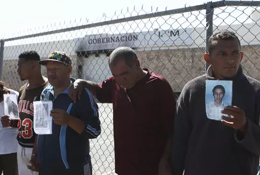 Migrants, mostly from Venezuela, hold photos of those who died in a fire at a Mexican immigration detention center, behind, during a prayer vigil outside the center in Ciudad Juarez, Mexico, April, 27, 2023. Four months after a fire in a Mexican immigration detention center at the border killed 40 migrants, some survivors are living in limbo at a Mexico City hotel, recovering from their injuries and awaiting the prosecution of their captors. (AP Photo/Christian Chavez, File)