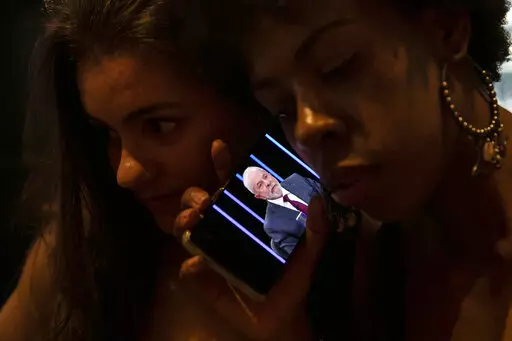 Women listen on a mobile phone to an interview of former president Luiz Inacio Lula da Silva, who is running again for president, with Jornal Nacional on TV Globo, outside a bar in Rio de Janeiro, Brazil, Thursday, August 25, 2022. The Superior Electoral Court, the country's top electoral authority, announced Thursday, Oct, 20, that it would be banning "false or seriously decontextualized" content that “affects the integrity of the electoral process.” No request from a prosecutor or complain