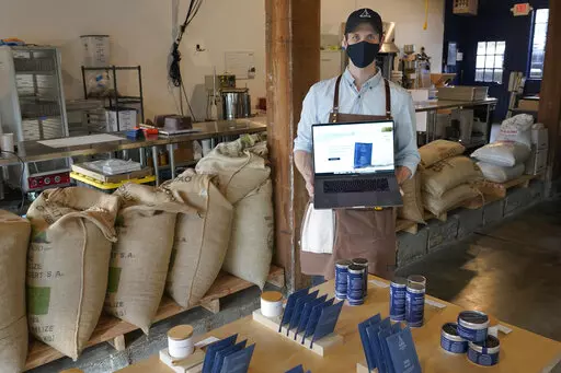 Kelly Van Arsdale poses for a photo Monday, Jan. 31, 2022, in Spinnaker Chocolate, the store he owns with his brother in Seattle where they make and sell single-origin chocolate bars. Van Arsdale is holding a laptop showing his company's website, and he says despite getting a lot of foot-traffic business, reaching customers online has been expensive and difficult. (AP Photo/Ted S. Warren)