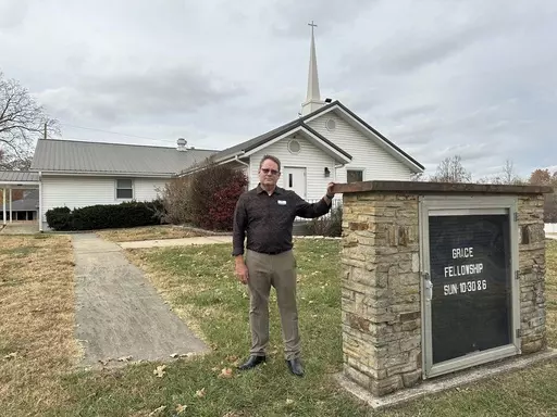 Pastor Kenny Batson stands near a sign displaying the worship service times of Grace Fellowship Church on Nov. 16, 2023, in El Dorado Springs, Mo. Batson was convicted of a series of crimes in the 1990s but became a Christian pastor after being released from prison. He was pardoned by Missouri Gov. Mike Parson. (AP Photo/David A.Lieb)